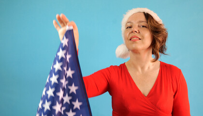 Wall Mural - A cheerful forty-year-old woman in a red sweater and a New Year's hat holds the US flag. Portrait of an adult woman on a blue background