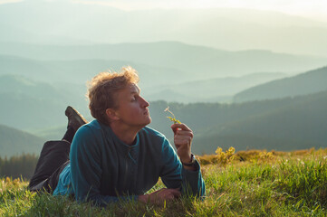 Wall Mural - A young man lies on the grass and smells a flower against the backdrop of mountains, rests in nature, enjoys his vacation, approaches, relaxation