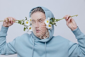 Wall Mural - a conceptual studio photograph of a man in a light blue hoodie, a mesh stretched over his face, standing on a light background, holding daisies near his face, looking into the camera