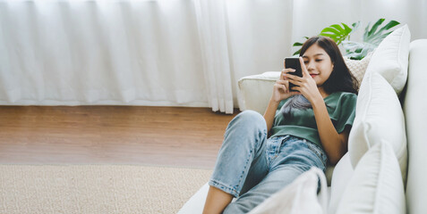 Wall Mural - Happy young asian woman relaxing at home. Female is lying down on sofa and using mobile smartphone