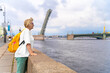 Blonde woman with backpack is standing on embankment, leaning on granite parapet, raised span of movable single-leafed bascule Trinity bridge in Saint Petersburg on background