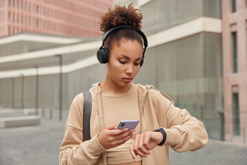 Serious curly haired woman focused at smartwatch to check pulse and results after cardio workout uses sport app on smartphone dressed in activewear listens music via headphones for vitality motivation