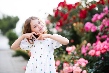 Poster - Happy smiling cheerful little child girl 6-7 year old show heart with fingers posing over rose flowers over nature background outside. Looking at camera. Cute sweet adorable kid having fun and play