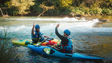 Two Smiled Caucasian Recreational Sportsmen Floating In Kayaks On The River, Giving High Five To Each Other, After Successful Descent Over Rapids