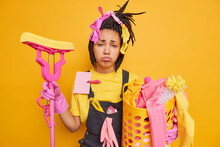 Exhausted Young Woman Tired After Doing Housework Holds Mop And Basket With Cleaning Tools And Laundry Has Busy Day Does Domestic Chores Looks Sadly At Camera Isolated Over Yellow Background