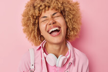 Overjoyed Young Woman With Curly Hair Laughs Out Happily Keeps Eyes Closed Hears Funny Story Happened With Friend Wears Casual Clothes Stereo Headphones Around Neck Isolated On Pink Background