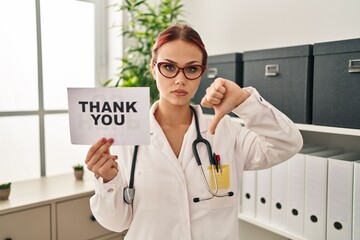 Canvas Print - Young caucasian woman wearing doctor uniform holding thank you banner with angry face, negative sign showing dislike with thumbs down, rejection concept