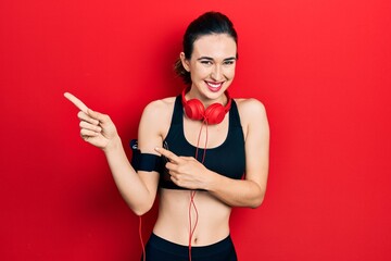 Young hispanic girl wearing gym clothes and using headphones smiling and looking at the camera pointing with two hands and fingers to the side.