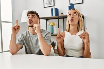 Poster - Young beautiful couple wearing casual clothes sitting on the table at home amazed and surprised looking up and pointing with fingers and raised arms.