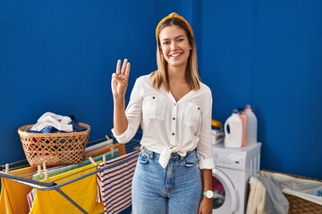 Sticker - Young blonde woman at laundry room showing and pointing up with fingers number three while smiling confident and happy.