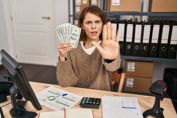 Canvas Print - Young beautiful woman working at small business ecommerce holding money with open hand doing stop sign with serious and confident expression, defense gesture