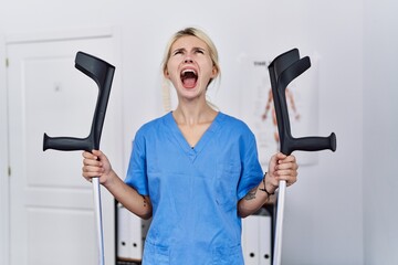 Sticker - Young physiotherapist woman holding crutches angry and mad screaming frustrated and furious, shouting with anger looking up.