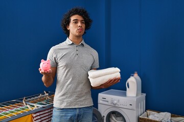 Poster - Hispanic man with curly hair holding clean towels and piggy bank looking at the camera blowing a kiss being lovely and sexy. love expression.