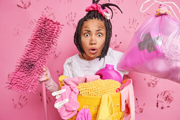 Domestic chores concept. Stunned young woman with braided hair holds dirty mop and bag of rubbish cannot believe own eyes does house cleaning stands near basin with laundry isolated on pink wall