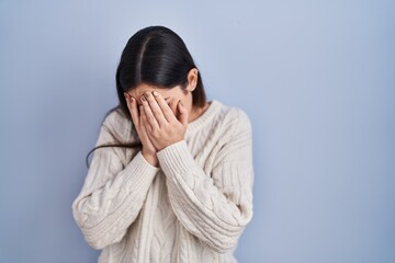 Poster - Young brunette woman standing over blue background with sad expression covering face with hands while crying. depression concept.