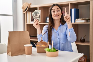 Sticker - Young brunette woman eating take away food at home holding money smiling with an idea or question pointing finger with happy face, number one