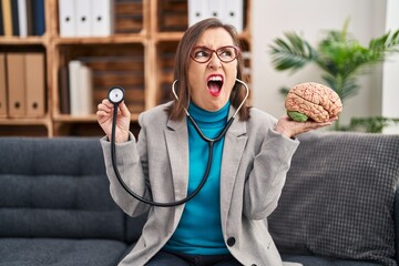 Wall Mural - Middle age hispanic woman working at therapy office holding brain angry and mad screaming frustrated and furious, shouting with anger. rage and aggressive concept.