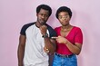 © Krakenimages.com - Young african american couple standing over pink background in hurry pointing to watch time, impatience, looking at the camera with relaxed expression