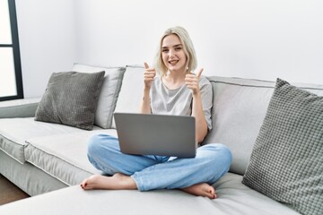 Poster - Young caucasian woman using laptop at home sitting on the sofa success sign doing positive gesture with hand, thumbs up smiling and happy. cheerful expression and winner gesture.