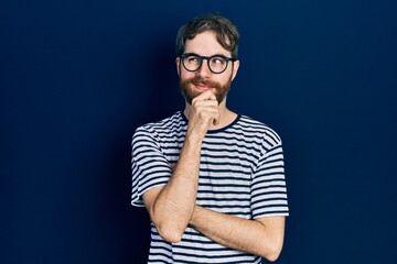 Wall Mural - Caucasian man with beard wearing striped t shirt and glasses with hand on chin thinking about question, pensive expression. smiling with thoughtful face. doubt concept.