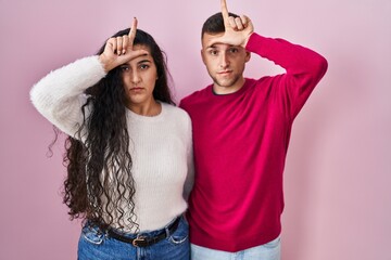 Sticker - Young hispanic couple standing over pink background making fun of people with fingers on forehead doing loser gesture mocking and insulting.