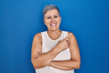 Poster - Middle age caucasian woman standing over blue background with a big smile on face, pointing with hand and finger to the side looking at the camera.