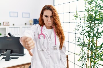 Poster - Young redhead woman wearing doctor uniform and stethoscope at the clinic looking unhappy and angry showing rejection and negative with thumbs down gesture. bad expression.