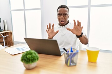 Poster - Young african man working at the office using computer laptop afraid and terrified with fear expression stop gesture with hands, shouting in shock. panic concept.