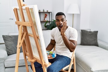 Canvas Print - Young african man painting on canvas at home afraid and shocked, surprise and amazed expression with hands on face