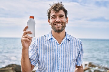 Sticker - Young handsome man holding sunscreen lotion looking positive and happy standing and smiling with a confident smile showing teeth