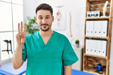 Wall Mural - Young physiotherapist man working at pain recovery clinic showing and pointing up with fingers number three while smiling confident and happy.