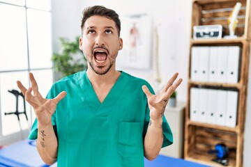 Canvas Print - Young physiotherapist man working at pain recovery clinic crazy and mad shouting and yelling with aggressive expression and arms raised. frustration concept.