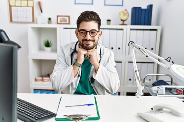 Sticker - Young man with beard wearing doctor uniform and stethoscope at the clinic laughing nervous and excited with hands on chin looking to the side