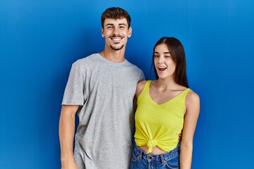 Poster - Young hispanic couple standing together over blue background winking looking at the camera with sexy expression, cheerful and happy face.