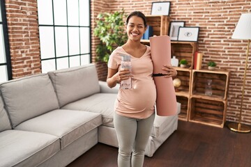 Sticker - Young pregnant woman holding yoga mat at home winking looking at the camera with sexy expression, cheerful and happy face.