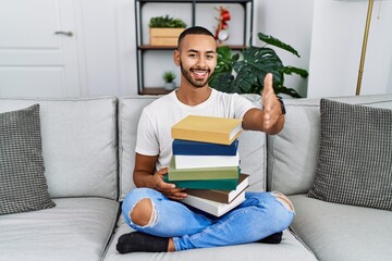 Poster - African american young man holding a pile of books sitting on the sofa smiling friendly offering handshake as greeting and welcoming. successful business.