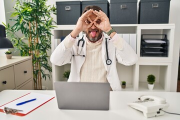 Poster - Handsome middle age doctor man working at the clinic doing ok gesture like binoculars sticking tongue out, eyes looking through fingers. crazy expression.