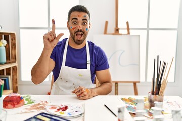 Poster - Young hispanic man with beard at art studio with painted face pointing finger up with successful idea. exited and happy. number one.