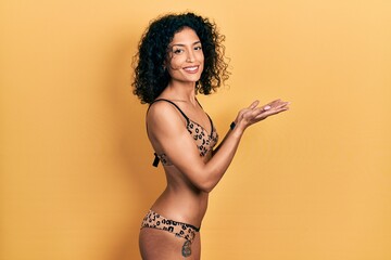 Young latin girl wearing bikini pointing aside with hands open palms showing copy space, presenting advertisement smiling excited happy