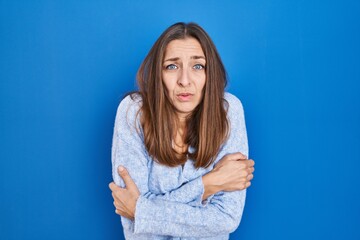 Young woman standing over blue background shaking and freezing for winter cold with sad and shock expression on face