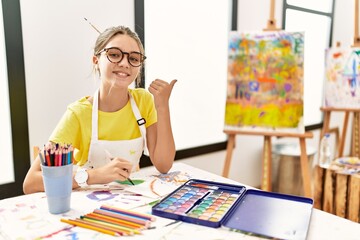 Sticker - Young brunette teenager at art studio pointing to the back behind with hand and thumbs up, smiling confident