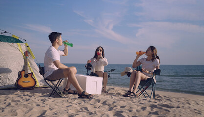 Group of happy Asian young friends sitting on camping chairs holding and drinking colored soft drinks on beach during summer vacation.