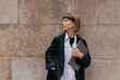 © PhotoBook - Charming female student with straight hair standing on the street. Pretty caucasian woman in black jacket and white t-shirt holding laptop and looking around.