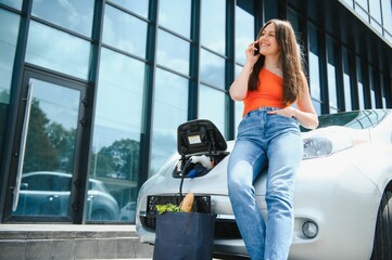 Wall Mural - Young woman is standing near the electric car and looks at the smart phone. The rental car is charging at the charging station for electric vehicles. Car sharing.