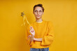 Playful woman holding magic wand and looking thoughtful while standing against yellow background