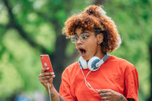 Surprised Girl In Headphones Looking At Mobile Phone Or Smartphone Outdoors