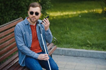 Visually impaired man with walking stick, sitting on bench in city park. Copy space