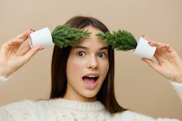 Wall Mural - a beautiful sweet woman stands on a light brown background in a white knitted sweater holding two small artificial Christmas trees in her hands, leans them against her face and smiles cheerfully