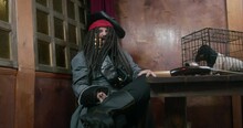 Old Pirate In Tricorn Hat And In Wig With Dreadlocks Crosses Legs And Rubs Sore Spot With Grimace On His Face. Walking Stick And Cage With Captive Dachshund Puppy On Table