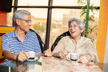 Happy indian senior couple sitting in living room with cup of tea or coffee, Retired elderly couple enjoying together at home, bonding and relationship. Aging 